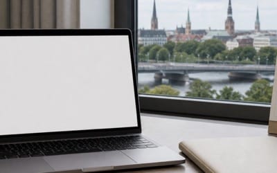 Rakstāmgalds ar klēpjdatoru, aizvērtu dokumentu mapi un kalendāru bez salasāmiem uzrakstiem; blakus krūze, aiz loga mierīga pilsētas aina, cilvēku attēlā nav.